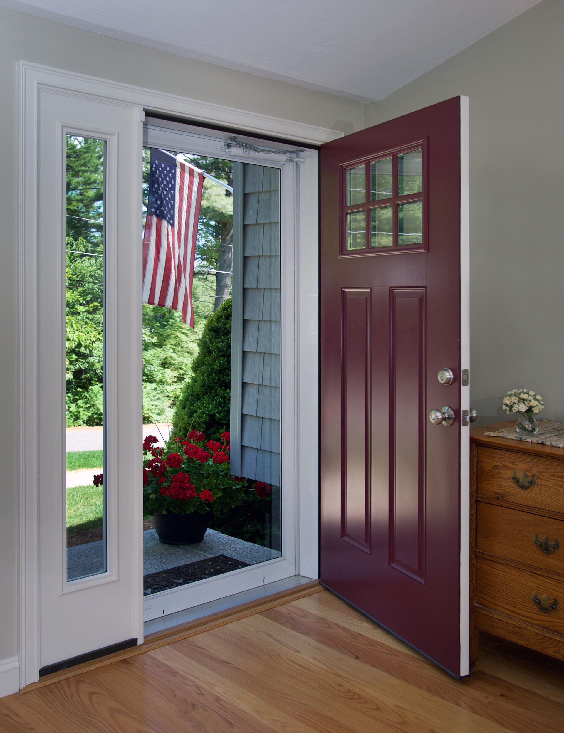 Open burgundy door with sidelight, revealing an American flag and greenery.