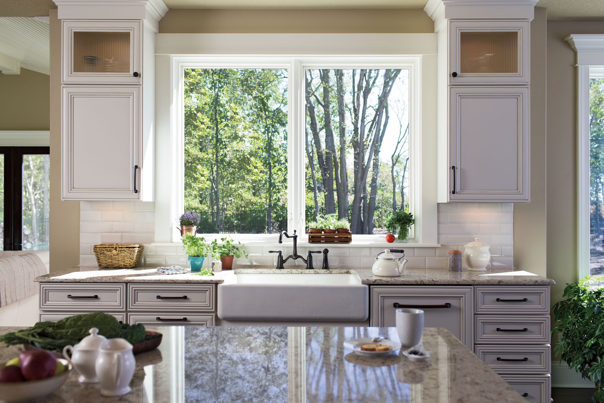 White kitchen with sink under large window, looking onto trees. Cabinets, countertop, and a teapot visible.