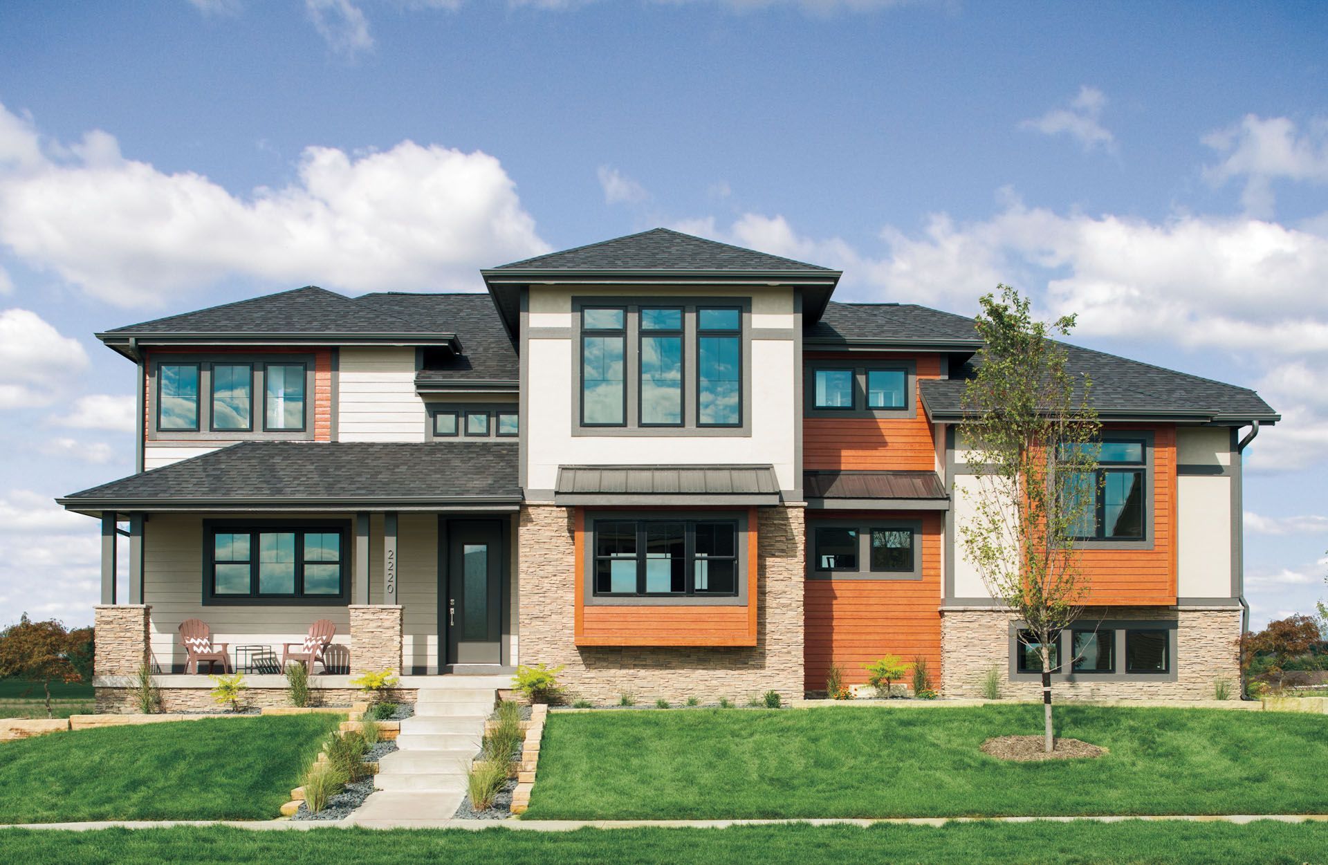 Modern two-story house with beige, orange, and stone exterior, black-framed windows, and green lawn against a blue sky.