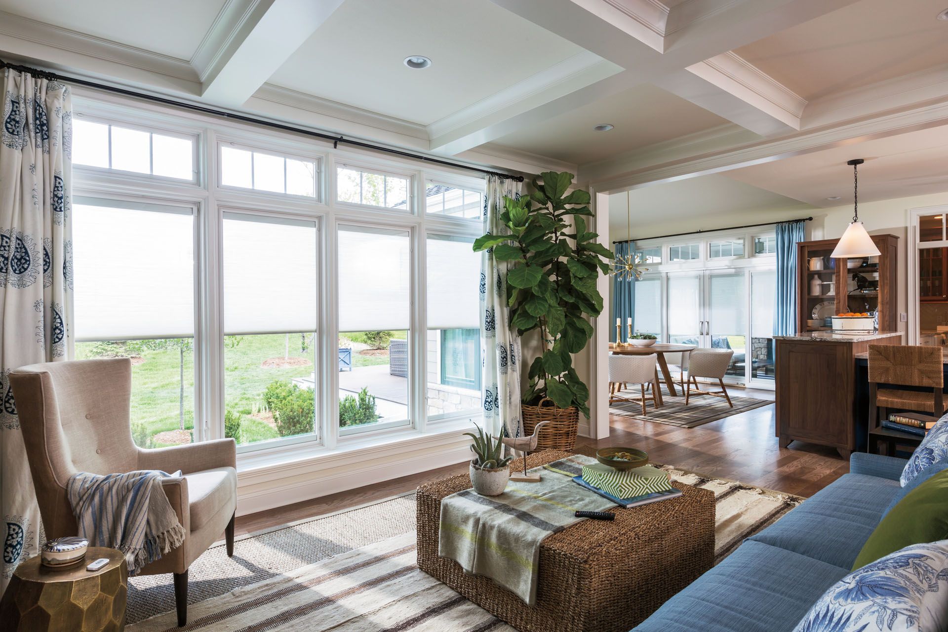 Living room with large windows, blue sofa, wicker coffee table, and tall plant.